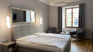 A modern hotel room with a large bed, a side table, a chair, a small table with a vase, and a window with curtains. - Hotel Gurmán (Horšovský Týn)