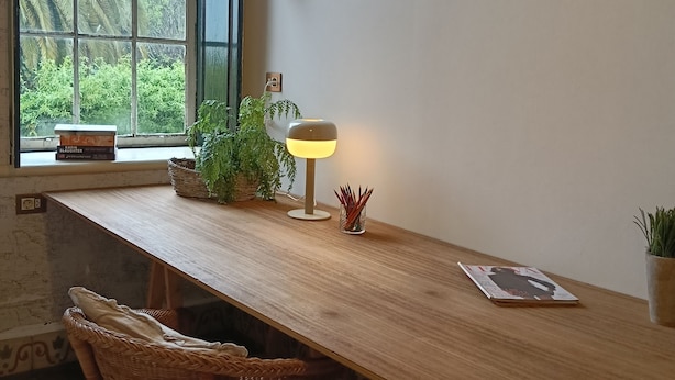 Un tavolo in legno con una lampada, una sedia in vimini e una finestra vista sul verde.
