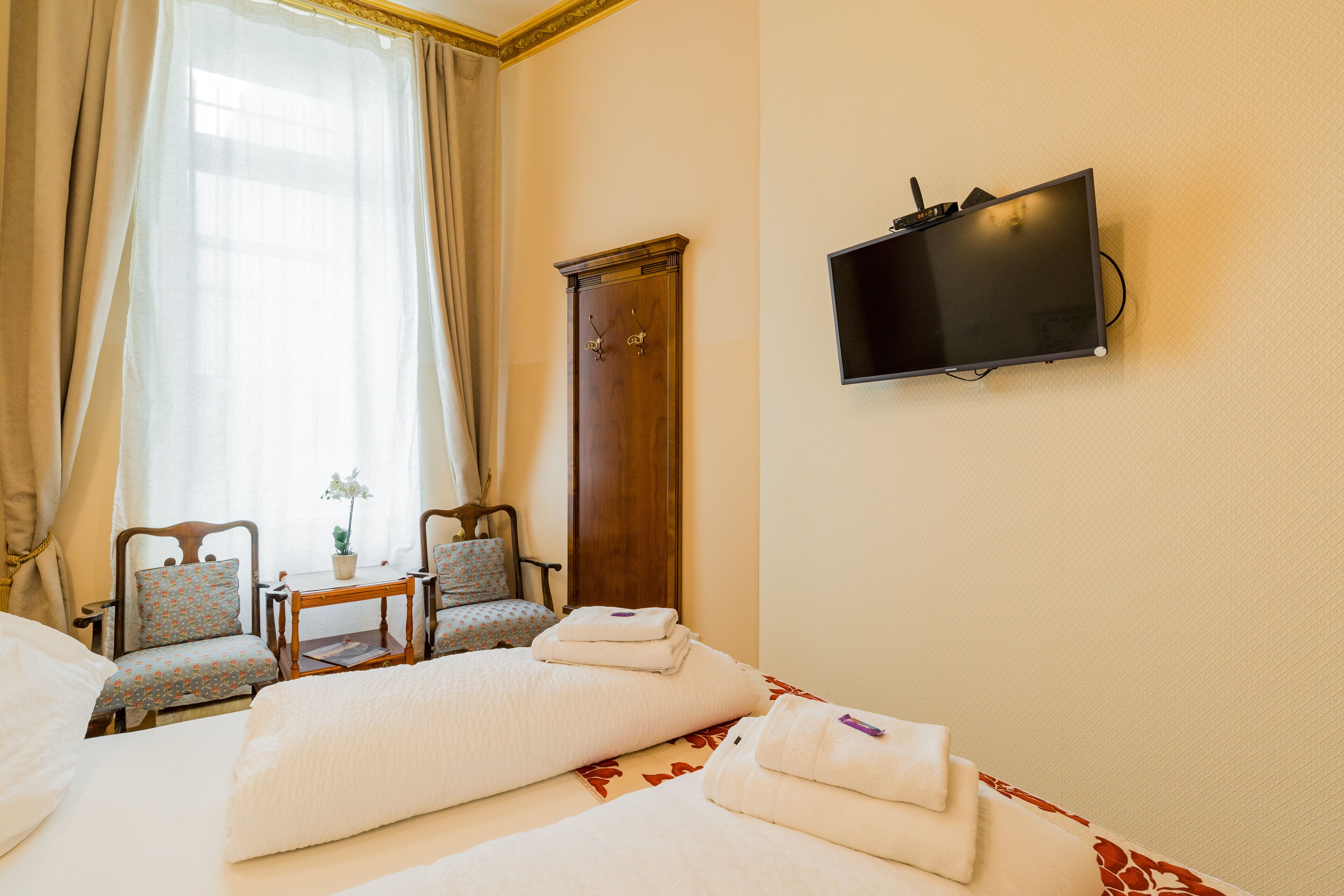 Quarto de hotel com duas camas, televisão, guarda-roupa de madeira e área de estar com cadeiras e mesa.