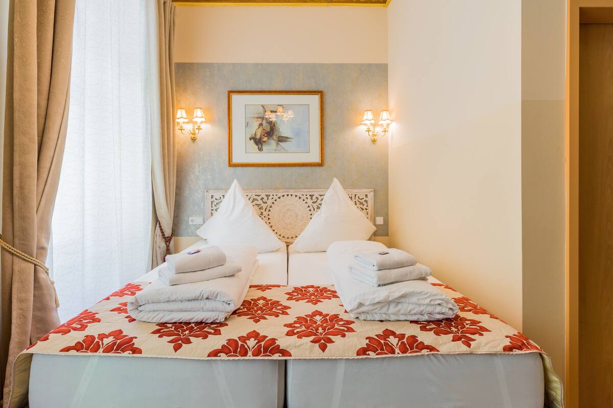A hotel room with a bed, two wall-mounted lamps, a framed artwork, and a window with curtains.