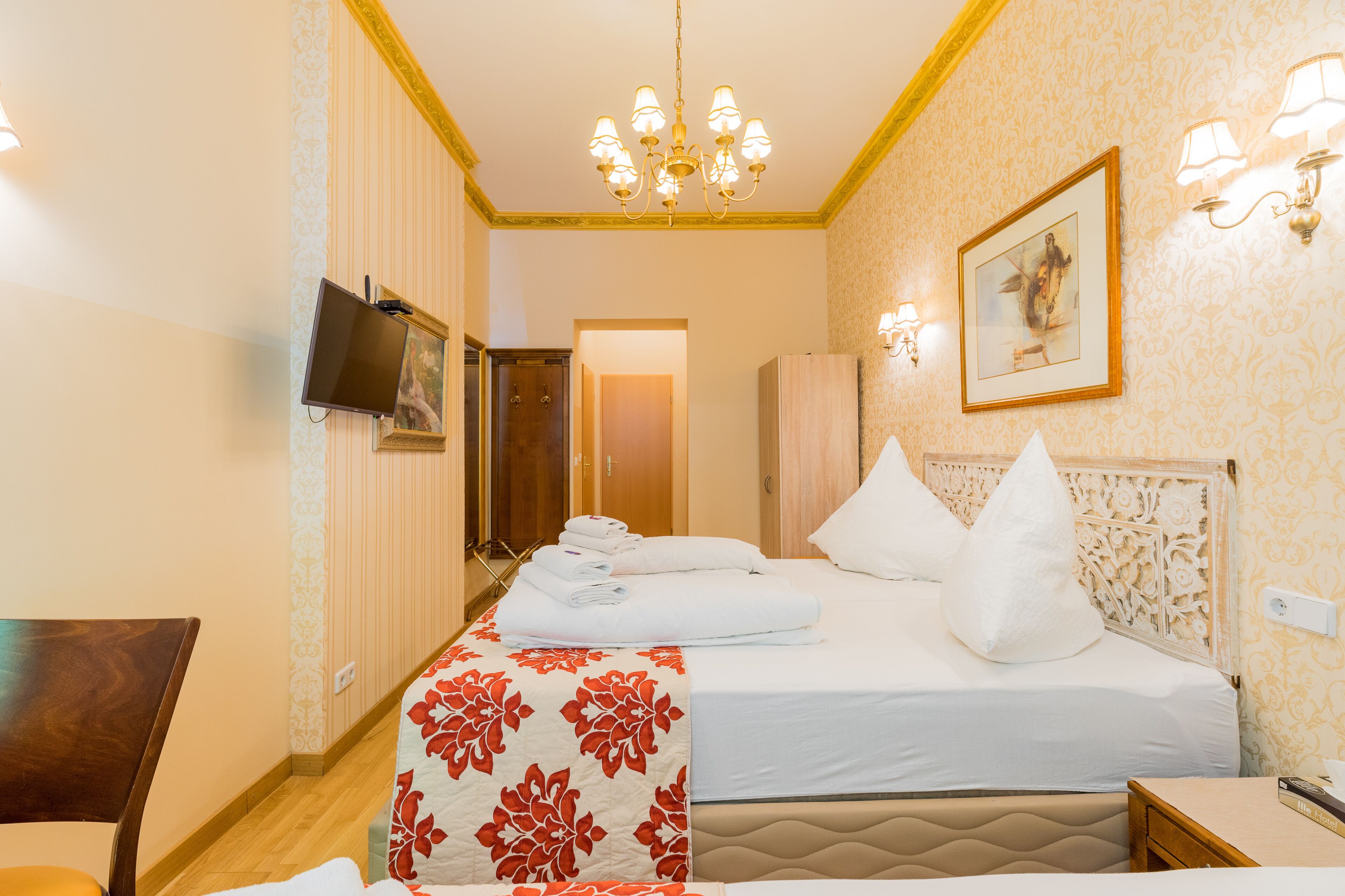 A hotel room with a bed, a chair, a desk, a television, and a chandelier.
