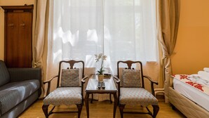 Una habitación con un armario de madera, un sofá, dos sillones estampados, una mesita con un florero y un control remoto, y una cama con colcha estampada.