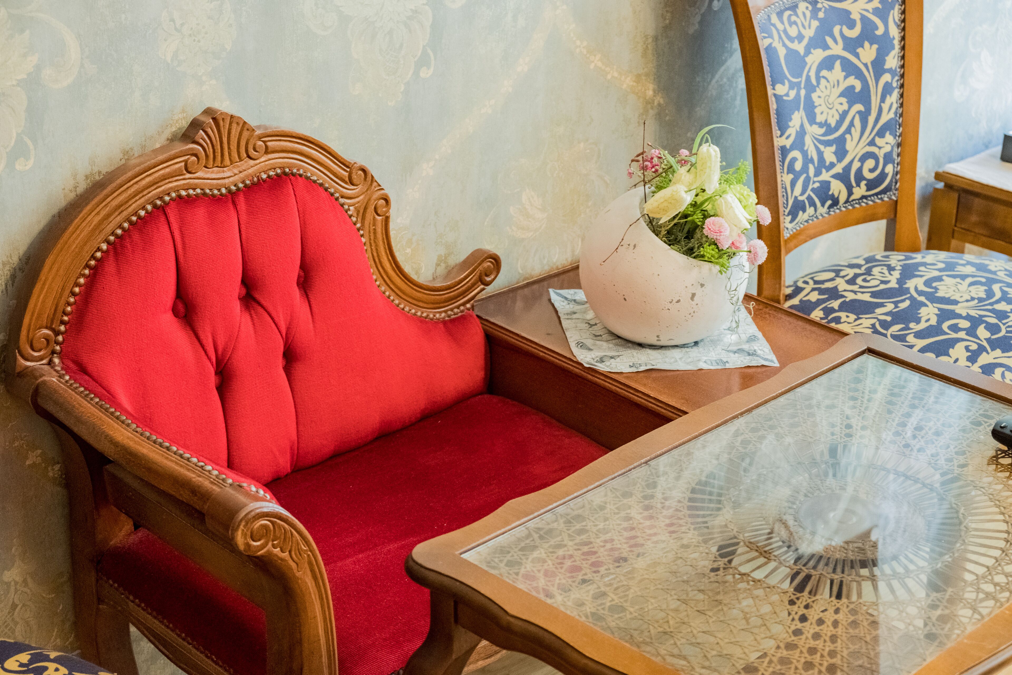 Un fauteuil rouge recouvert de tissu, avec une structure en bois, une table en verre ornée d’un motif en dentelle et un vase rempli de fleurs.