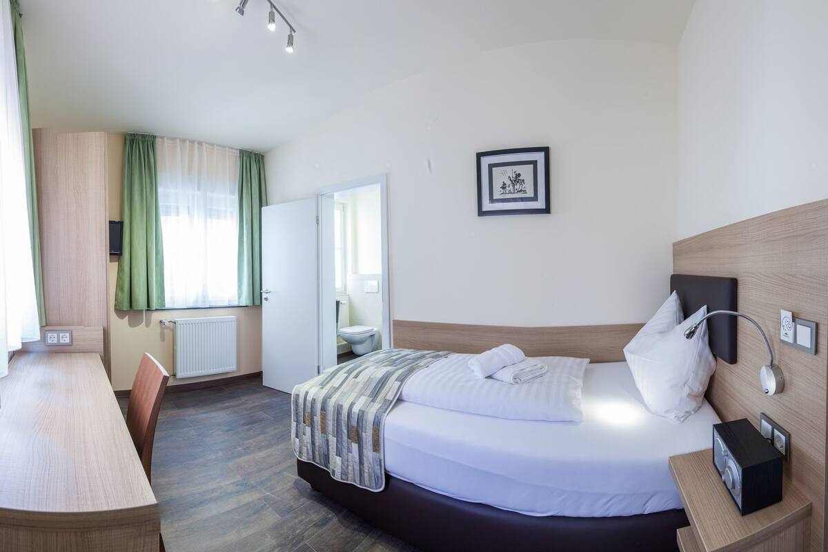 A hotel room with a bed, bedside table, and a desk with a chair. There is a window with curtains, a radiator, and a framed picture on the wall.