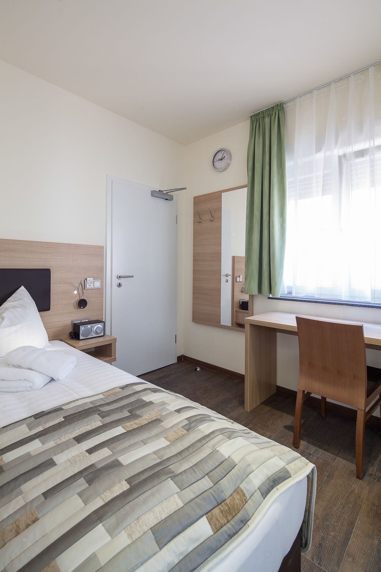 Habitación de hotel con cama, escritorio, silla, espejo, reloj y ventana con cortinas.
