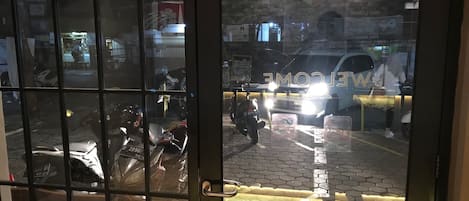 A view through a glass door with a 'WELCOME' sign, a motorcycle parked outside, and a reflection of a vehicle on the glass.