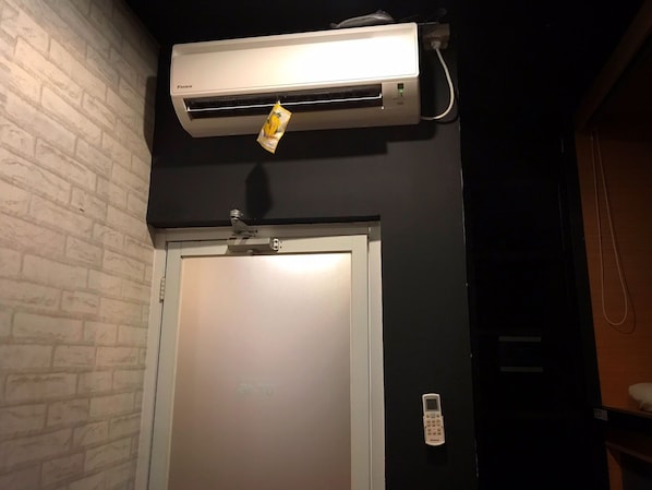 A room with a white air conditioner mounted on a brick wall, a door with a handle, and a small white device on the wall. - Tokyo Cubo - Hostel (Bandung)