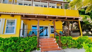 Un edificio amarillo de dos pisos con un porche cubierto, una escalera que conduce a la entrada y vista al mar.