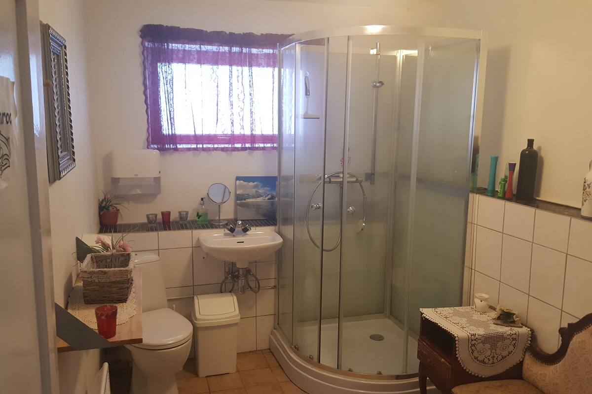 Un baño con ducha envuelta en vidrio, un lavamanos, un inodoro y una ventana con cortina.