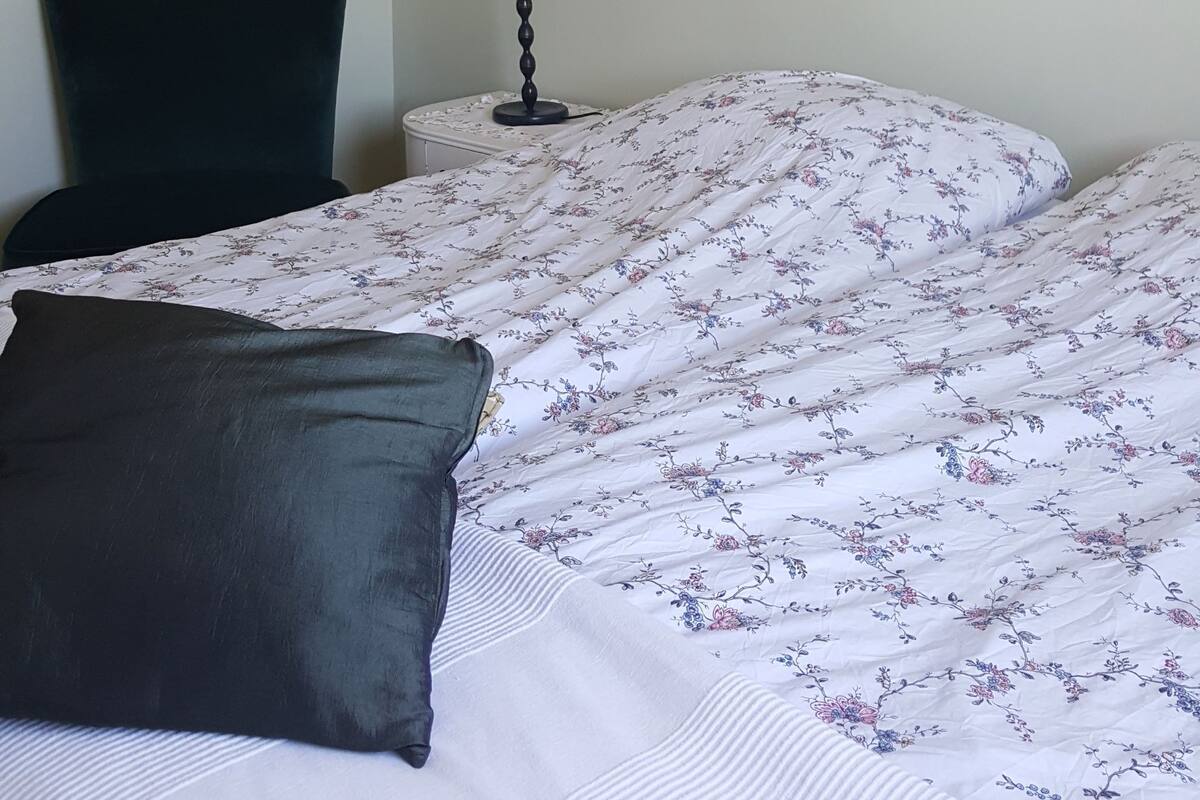 Una cama bien hecha con ropa de cama floral, una silla oscura, una mesita de noche con una lámpara y un estante con objetos decorativos.