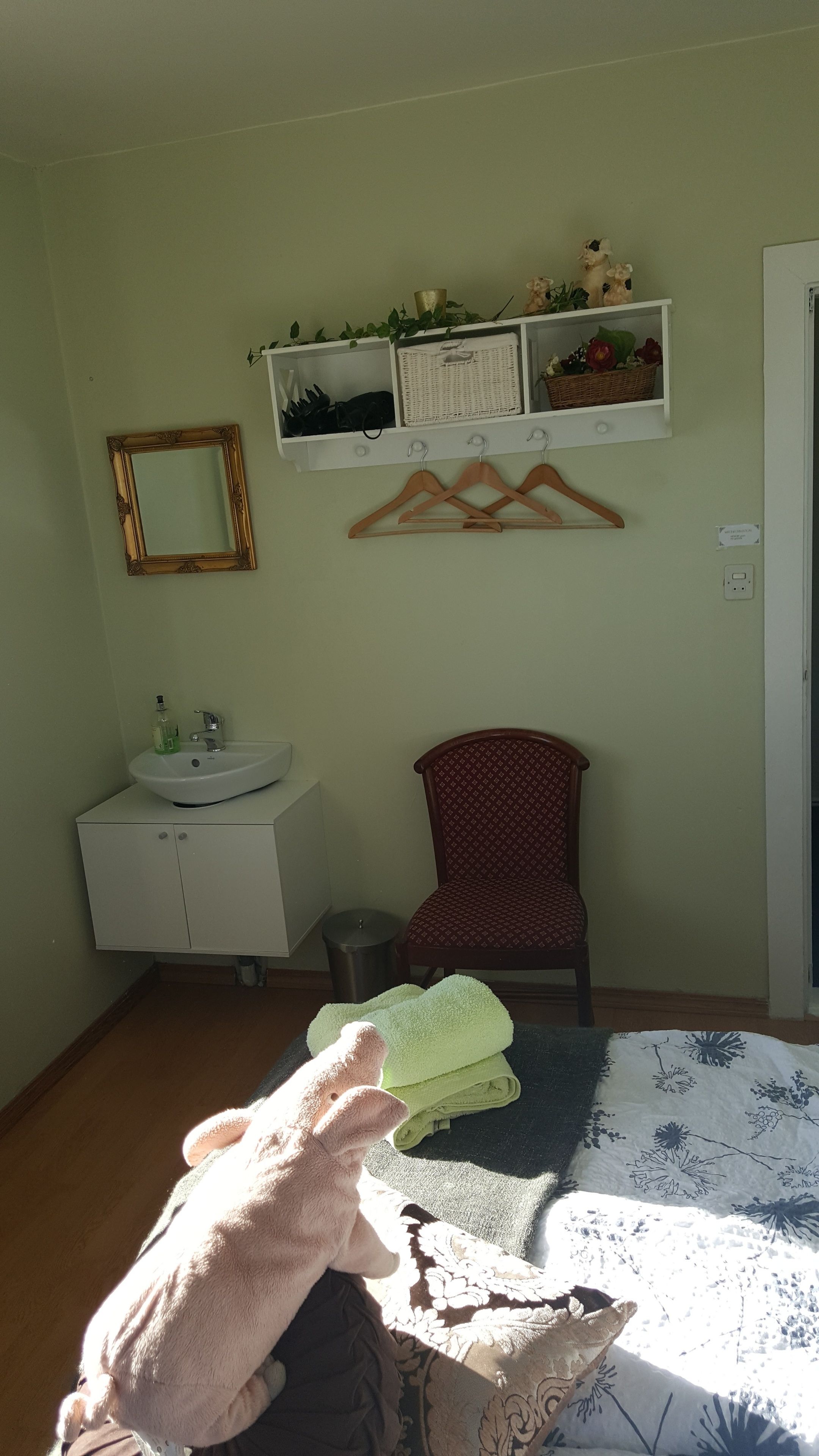 Quarto com pia branca, espelho, cadeira, cama com colcha estampada e prateleira com objetos decorativos.