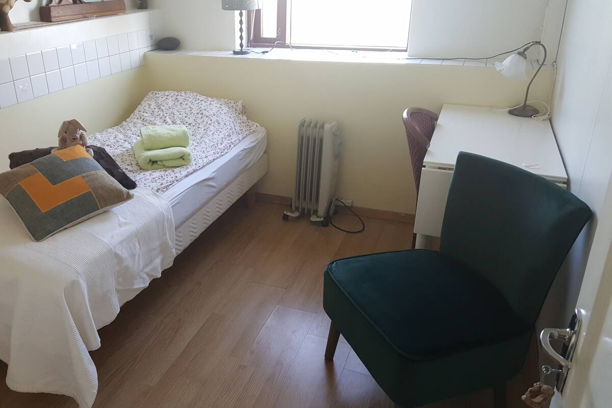 Una habitación pequeña y limpia con cama, silla, radiador y ventana.