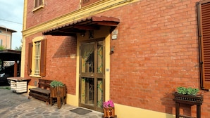 A red brick building with a yellow awning, a wooden bench, and a planter with flowers. - B&B Il Cantastorie Casa Molinari-Boldrini (Castelfranco Emilia)