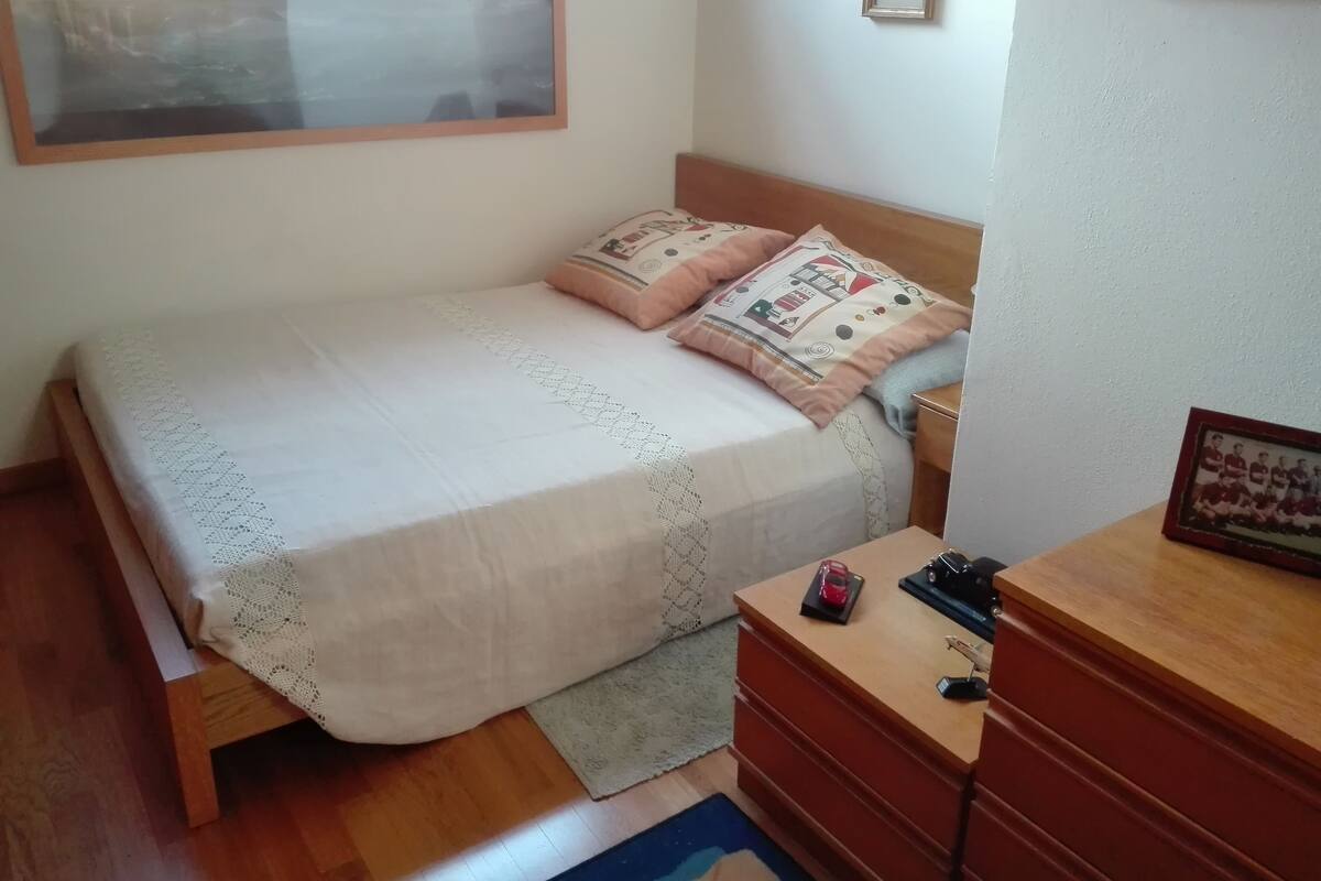 A bedroom with a bed, a wooden dresser, a nightstand, a framed picture, and a window with a view.