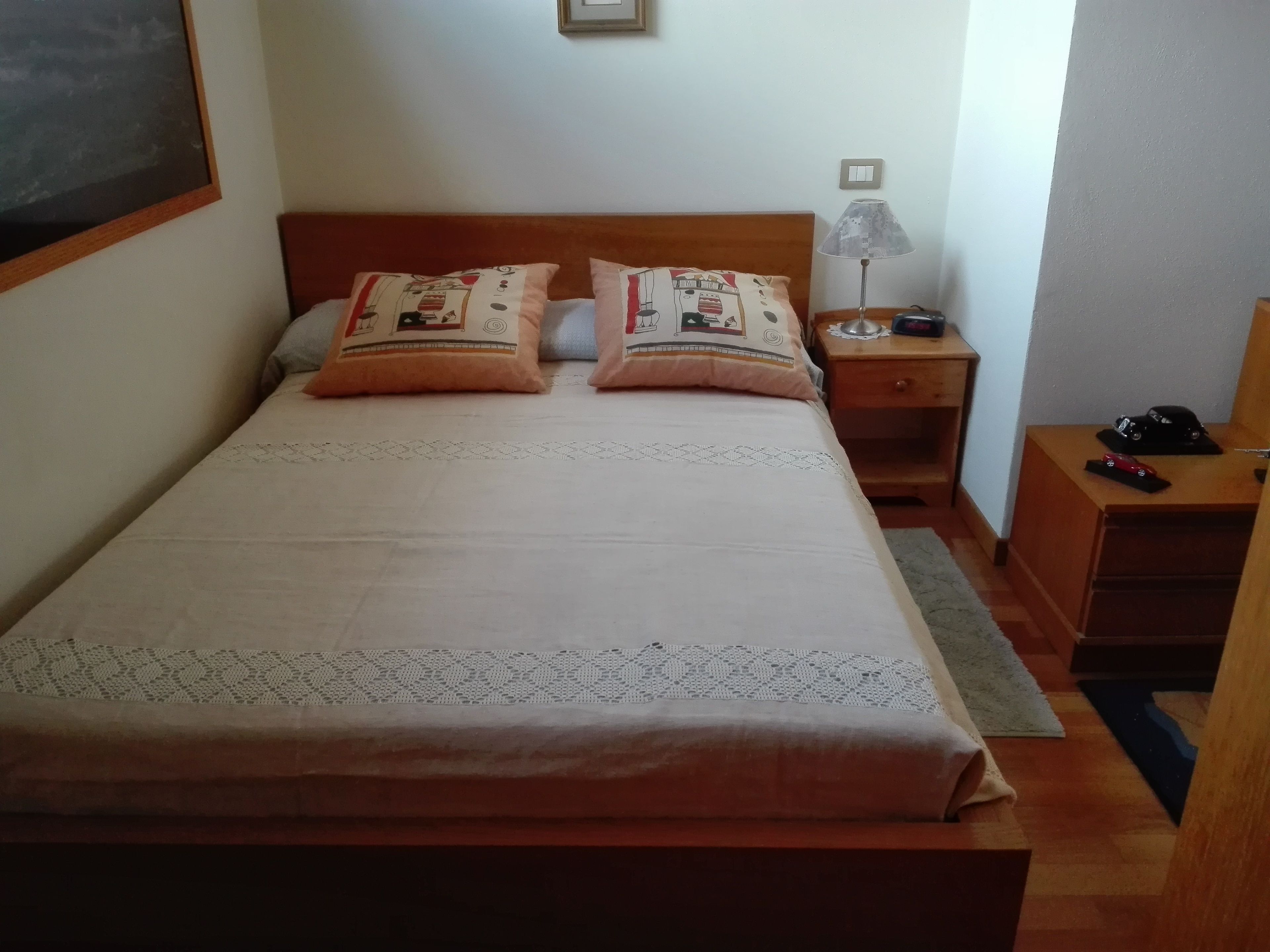 Una cama bien hecha con almohadas estampadas, una mesita de noche con lámpara y una pequeña cómoda.