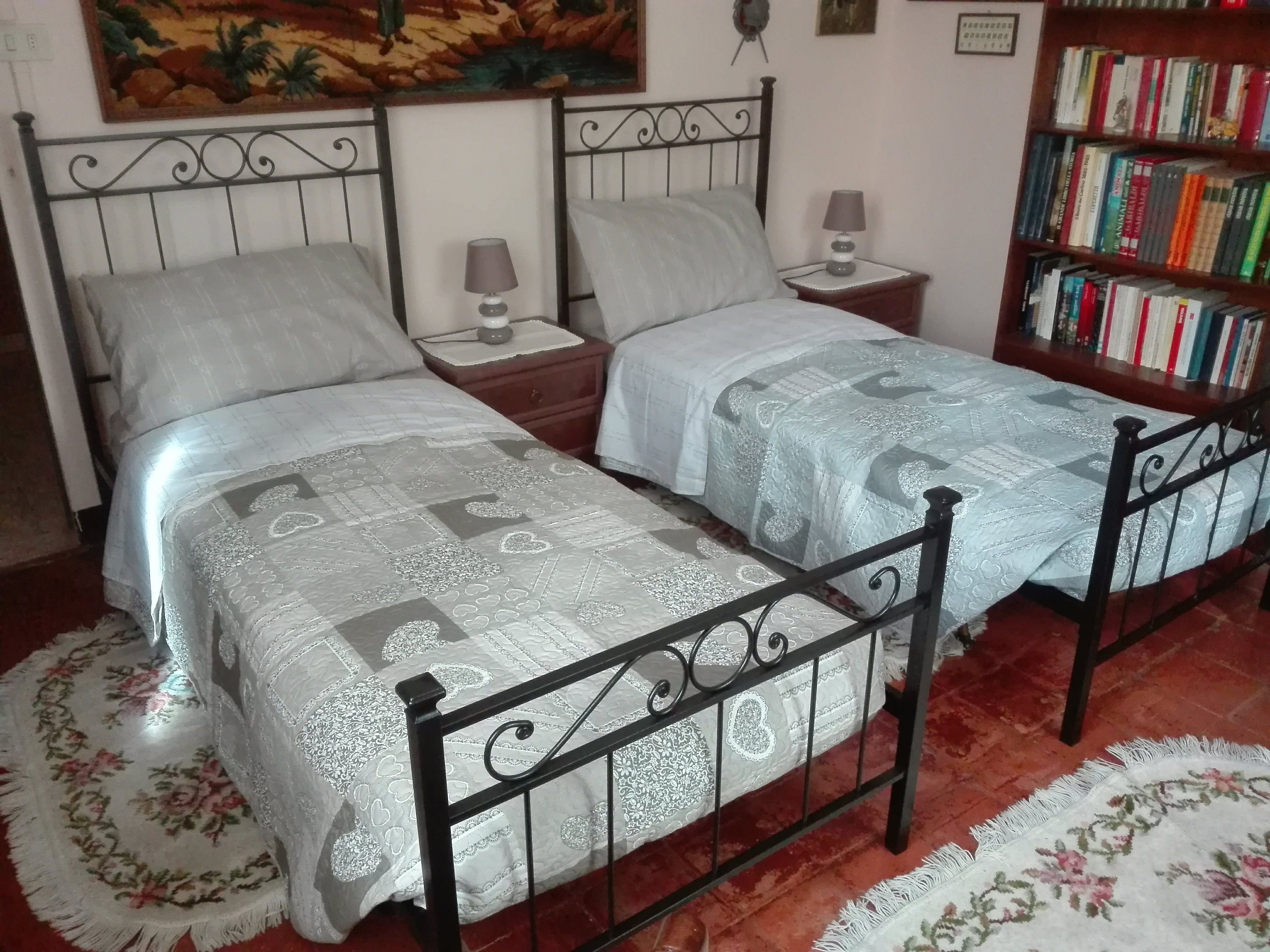 Een tweepersoonsbed met een decoratieve sprei, twee nachtkastjes met lampen, een boekenplank en een bloemenkleed.