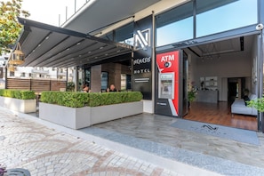 Fachada de hotel moderna com área de estar coberta ao ar livre, um caixa eletrônico e uma recepção.