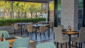 An outdoor cafe with tables and chairs, a brick wall, and a view of trees and a parked van. - Nemesis Hotel (Sarandë)