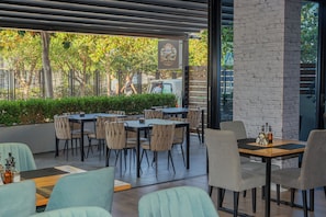 Un café al aire libre con mesas y sillas, un muro de ladrillo y vistas a árboles y una furgoneta estacionada.