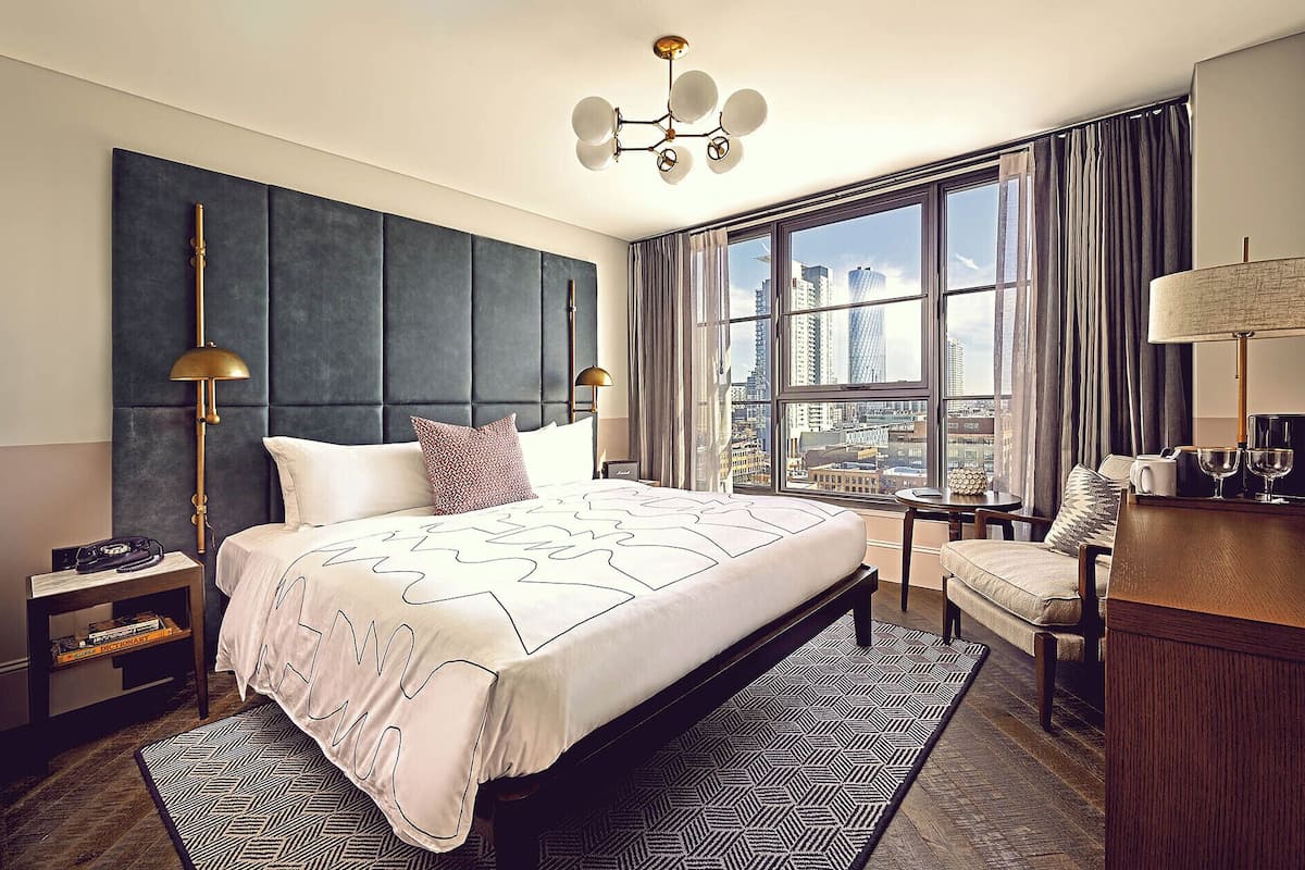 A modern hotel room with a large bed, a city view, a desk with a lamp, and a chair.