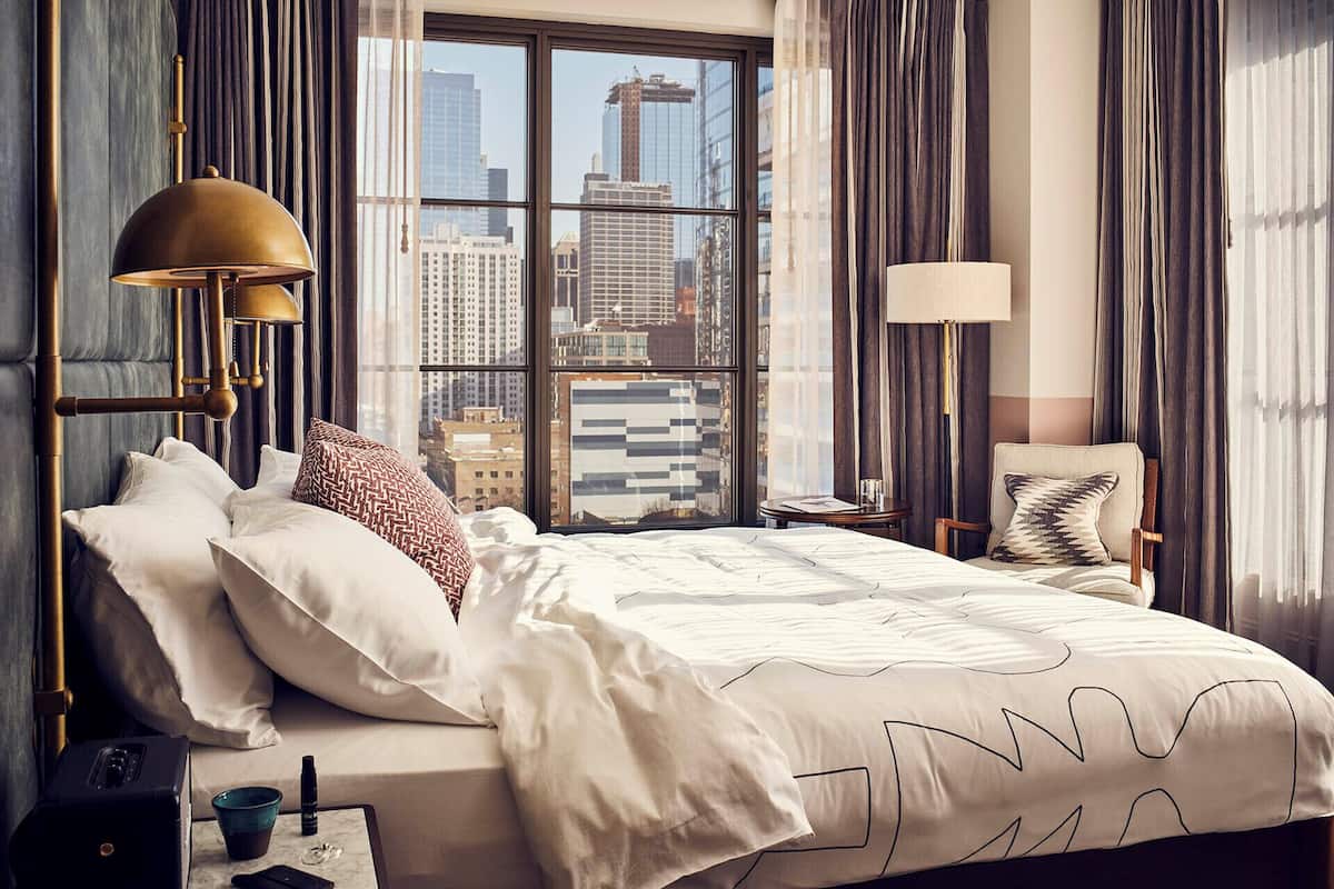 A hotel room with a large bed, two bedside lamps, a view of the cityscape through large windows, and a city skyline visible outside.