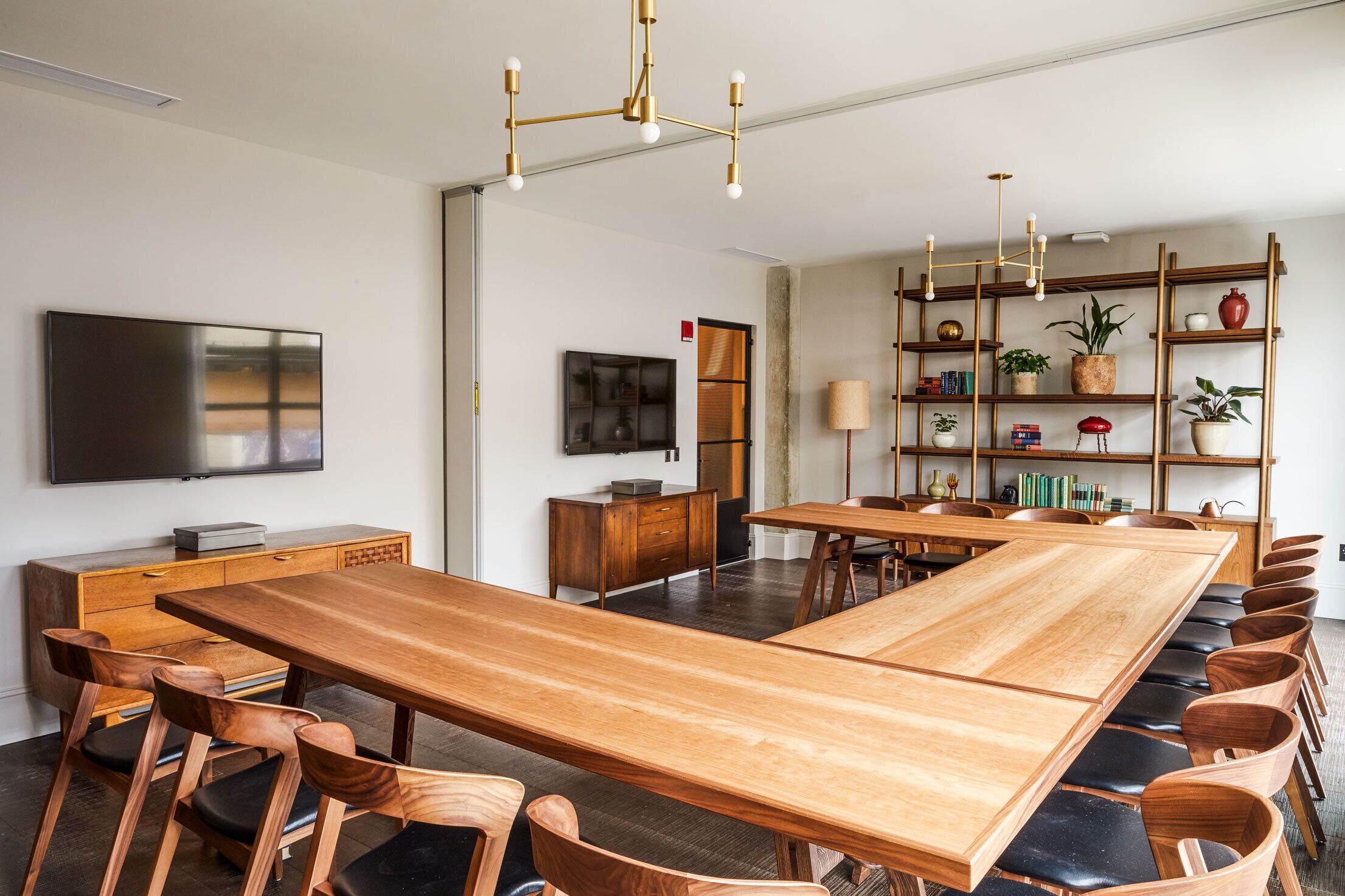 Un comedor amplio con una larga mesa de madera y sillas, un televisor fijado a la pared y una estantería con diversos objetos.