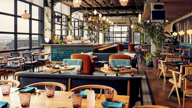 A modern restaurant with high ceilings, exposed beams, and large windows. Tables are set with blue napkins and pink glassware. - The Hoxton, Chicago (Chicago)