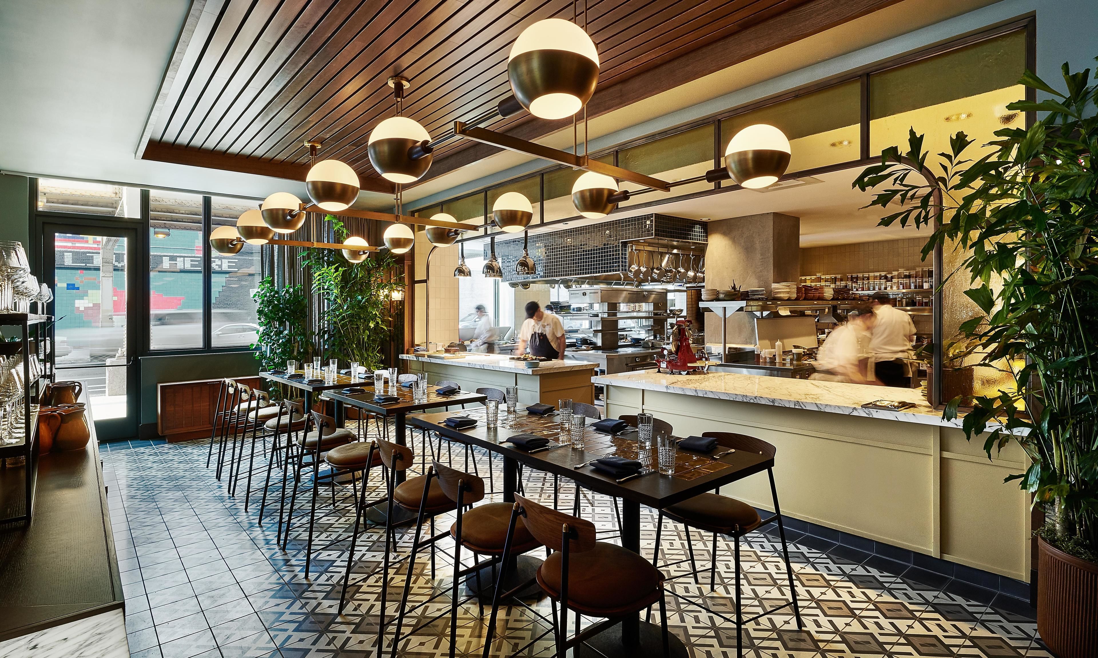 A restaurant interior with a long counter, multiple tables, and chairs. There are hanging pendant lights, a tiled floor, and potted plants.