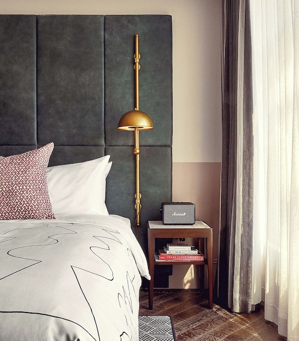 A neatly arranged bedroom with a tufted headboard, a bedside lamp, a small table with books, and a bed with patterned bedding.
