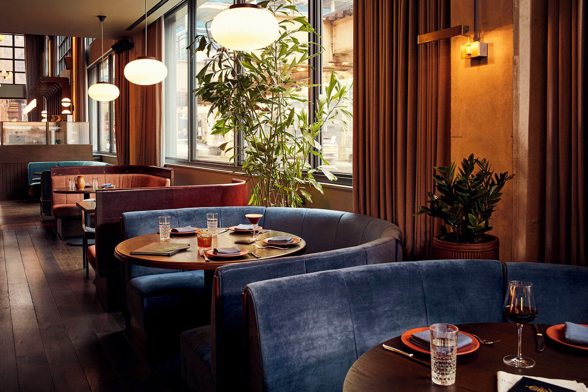 A restaurant interior with round tables, blue upholstered booths, and a large window with curtains.
