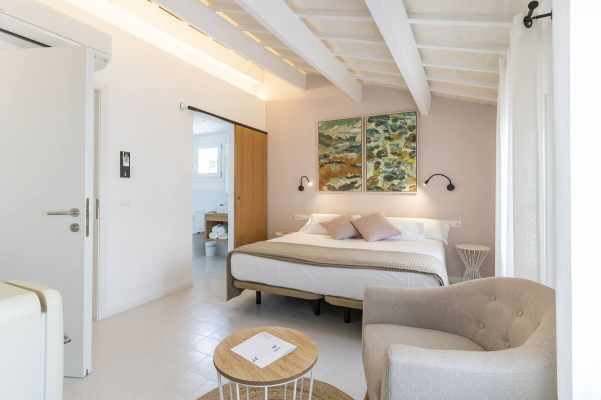 A bedroom with a bed, a chair, a small table, and artwork on the wall.