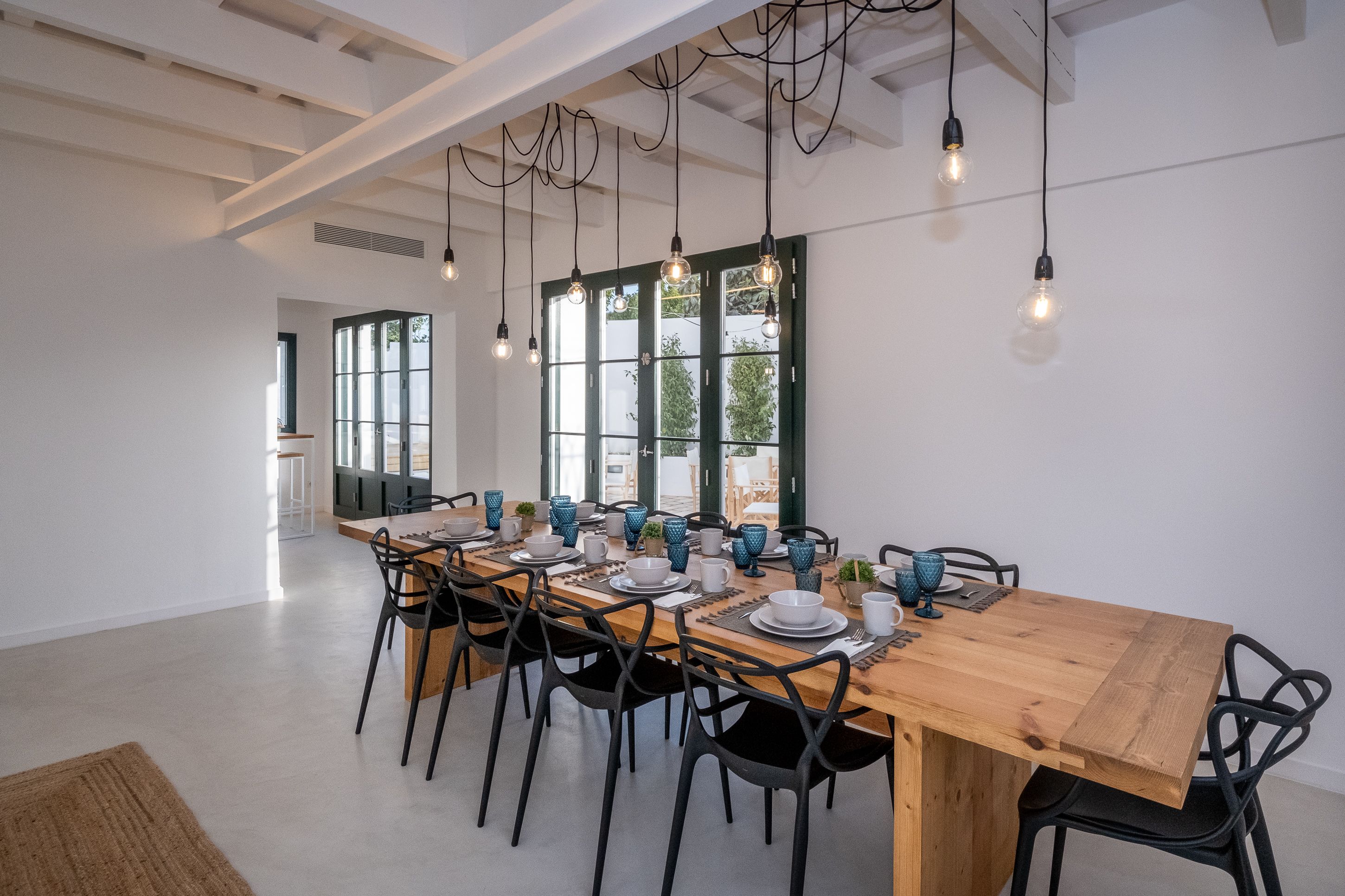 Comedor con una mesa de madera alargada dispuesta para seis comensales, sillas negras y lámparas colgantes.