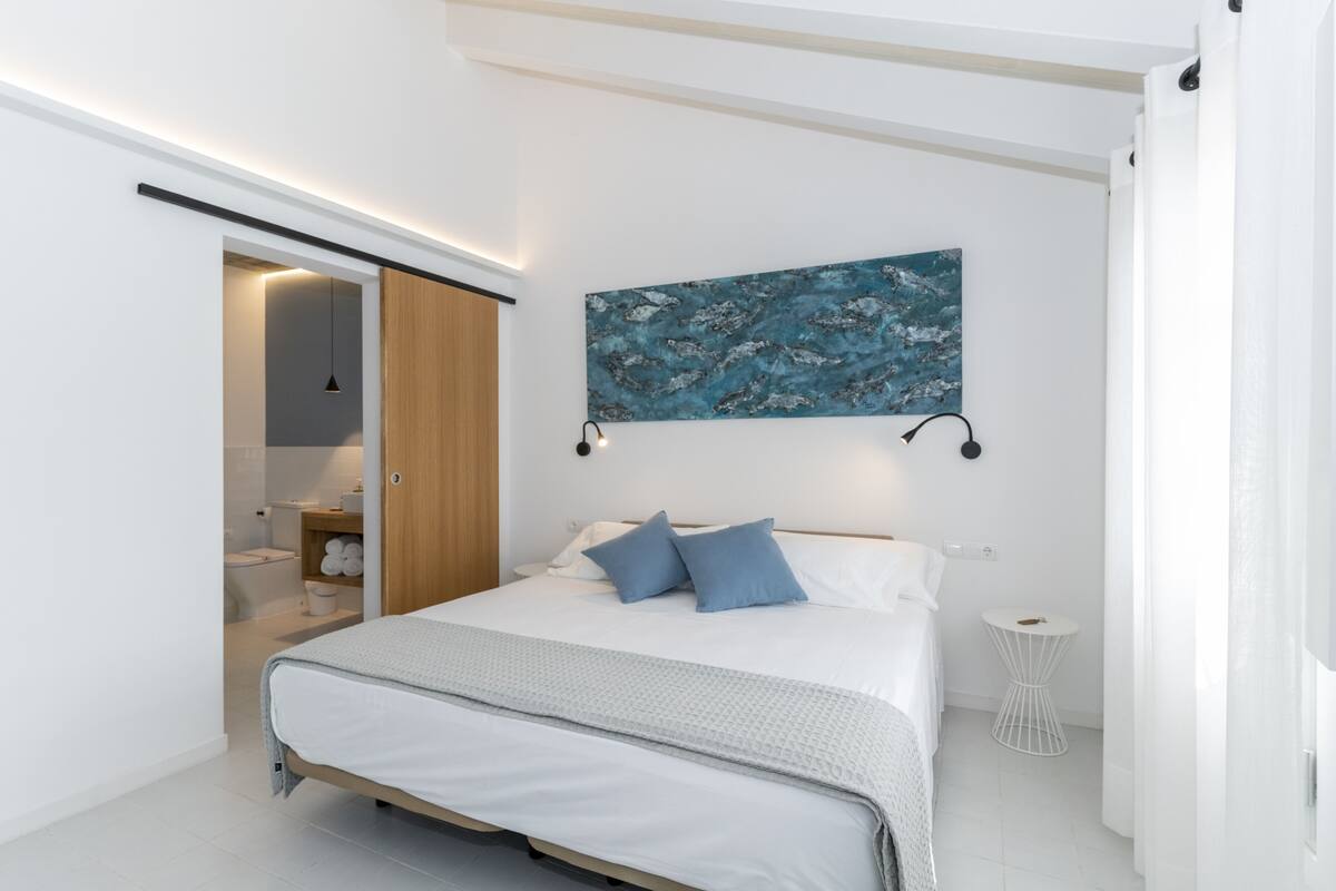 A modern bedroom with a bed, a painting, a bedside table, and a bathroom visible through an open door.