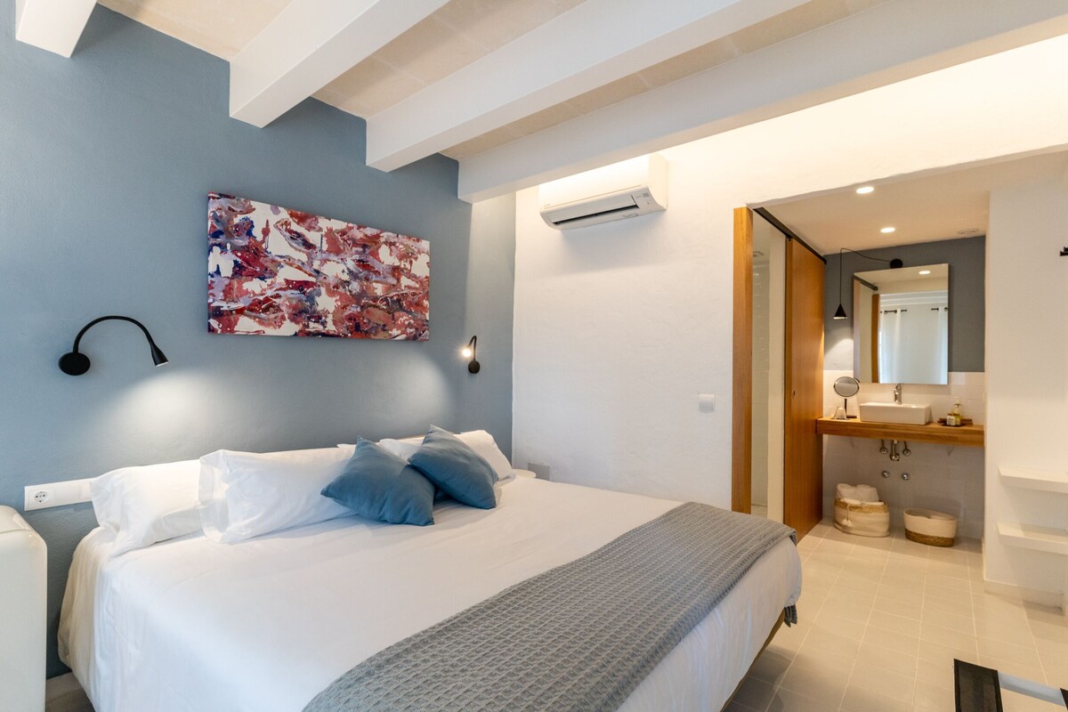 A modern bedroom with a bed, bedside lamps, a wall-mounted air conditioner, and a bathroom with a sink and mirror.