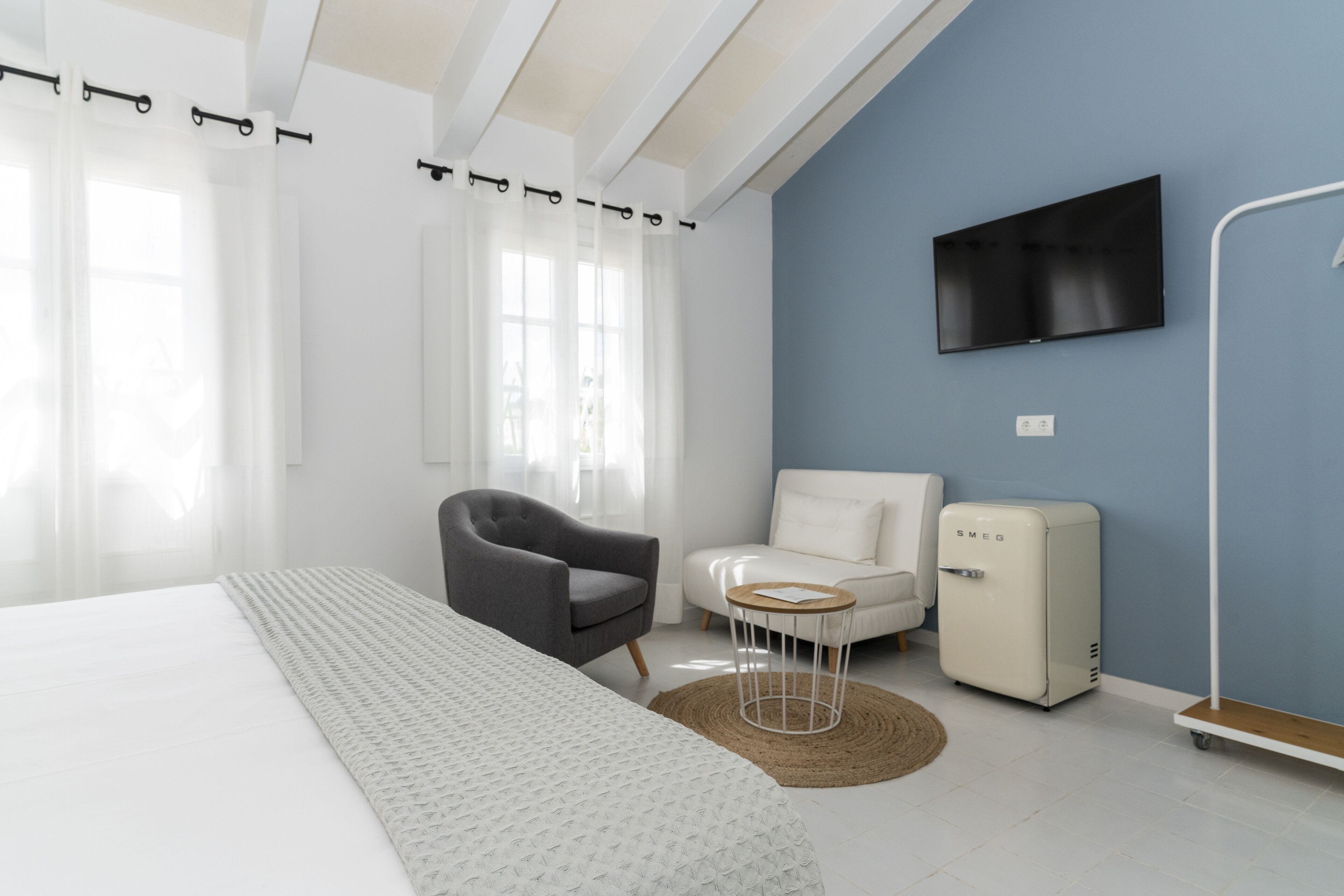 Habitación de hotel moderna con una cama grande, un sillón gris, un sillón blanco, una mesita blanca, un refrigerador blanco y un televisor montado.