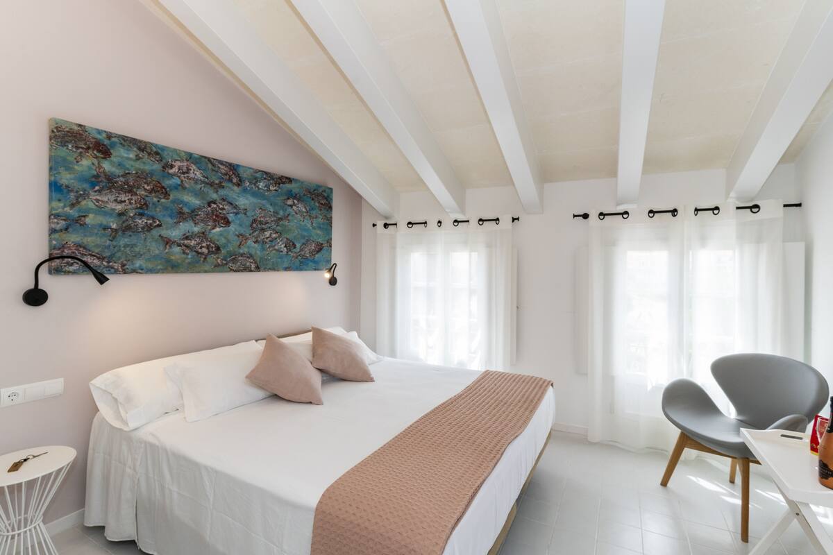 A bedroom with a bed, a chair, a table, a painting, and a window with curtains.