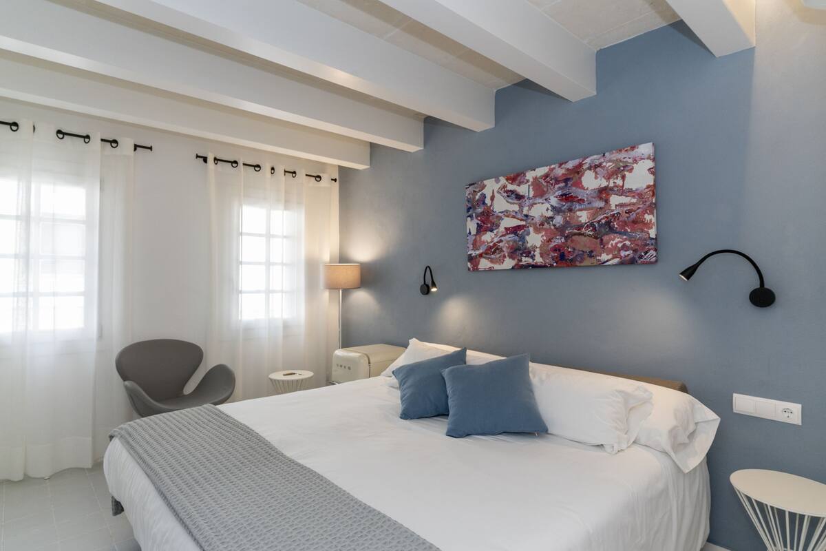 A modern bedroom with a large bed, a chair, a bedside table, and a wall-mounted lamp.