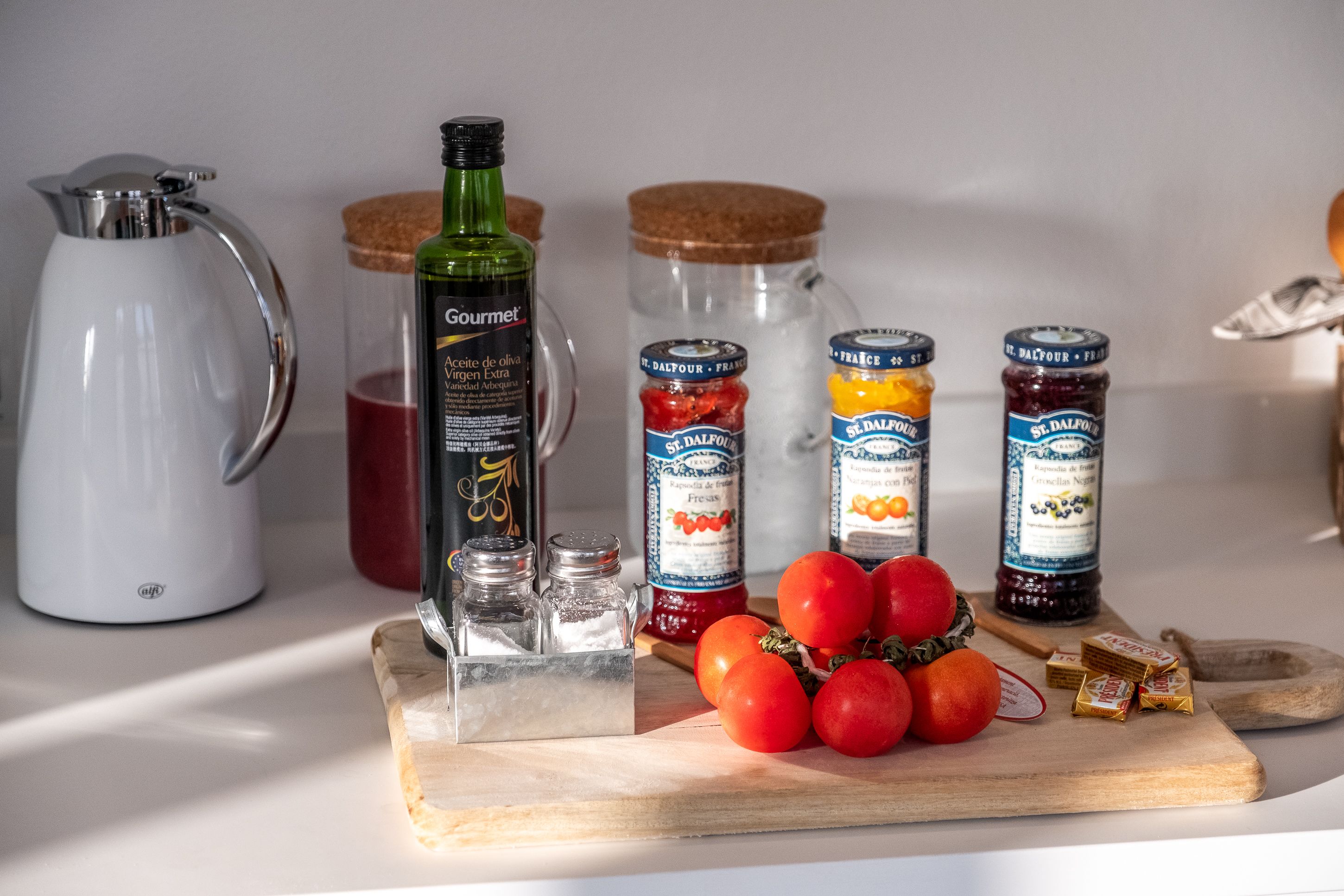 A kitchen counter with a coffee maker, a bottle of olive oil, jars of jam, and fresh tomatoes.