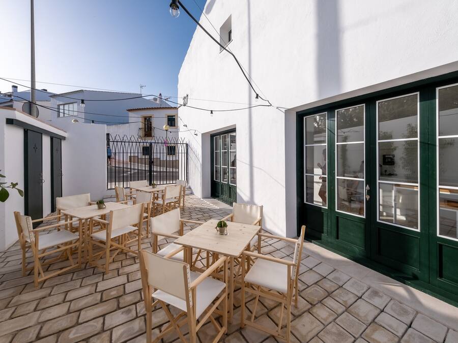 An outdoor patio with wooden tables and chairs, a small plant, and a white building with green-framed windows.