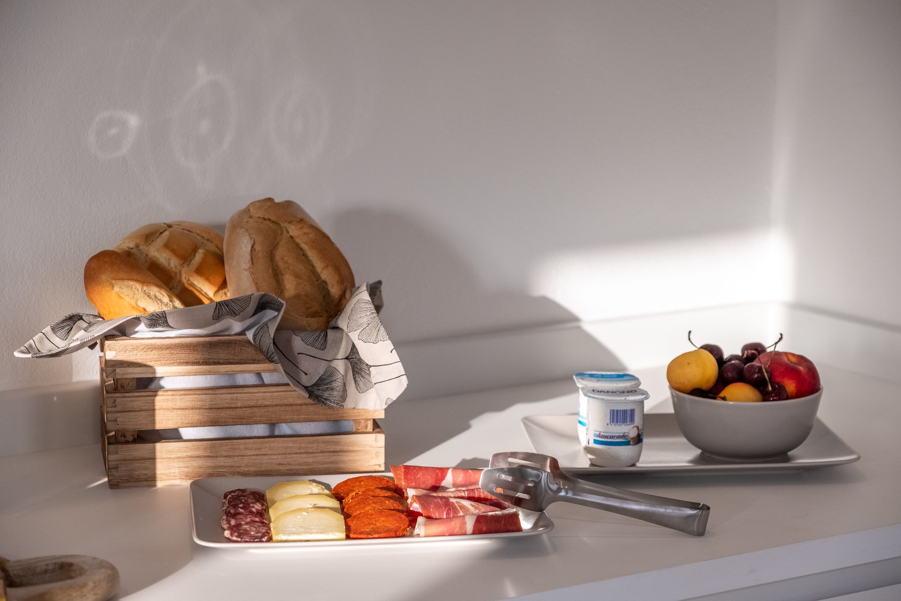 Desayuno con pan, queso, embutidos, fruta y yogur sobre una mesa.