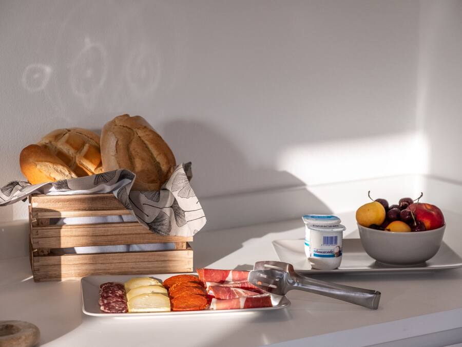 Desayuno con pan, queso, embutidos, fruta y yogur sobre una mesa.