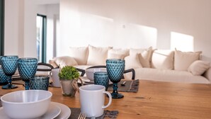 Un coin repas avec une table en bois, agrémentée de verres bleus, d’assiettes blanches et d’une tasse blanche. Une petite plante en pot est posée sur la table, et un canapé avec des coussins se trouve à l’arrière-plan.
