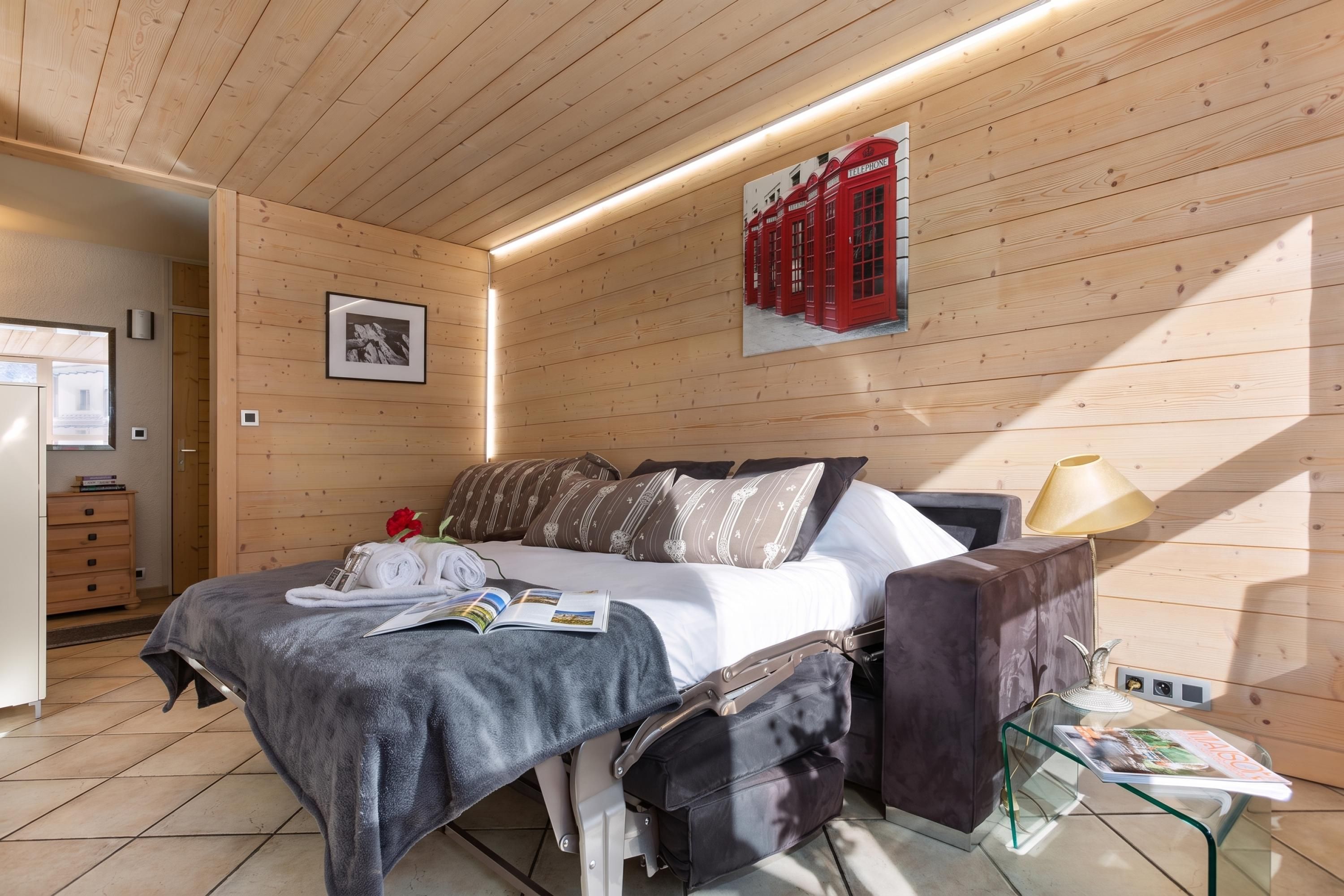 A modern bedroom with a bed, pillows, a bedside table with a lamp, a glass table with a magazine, and a wall-mounted picture.