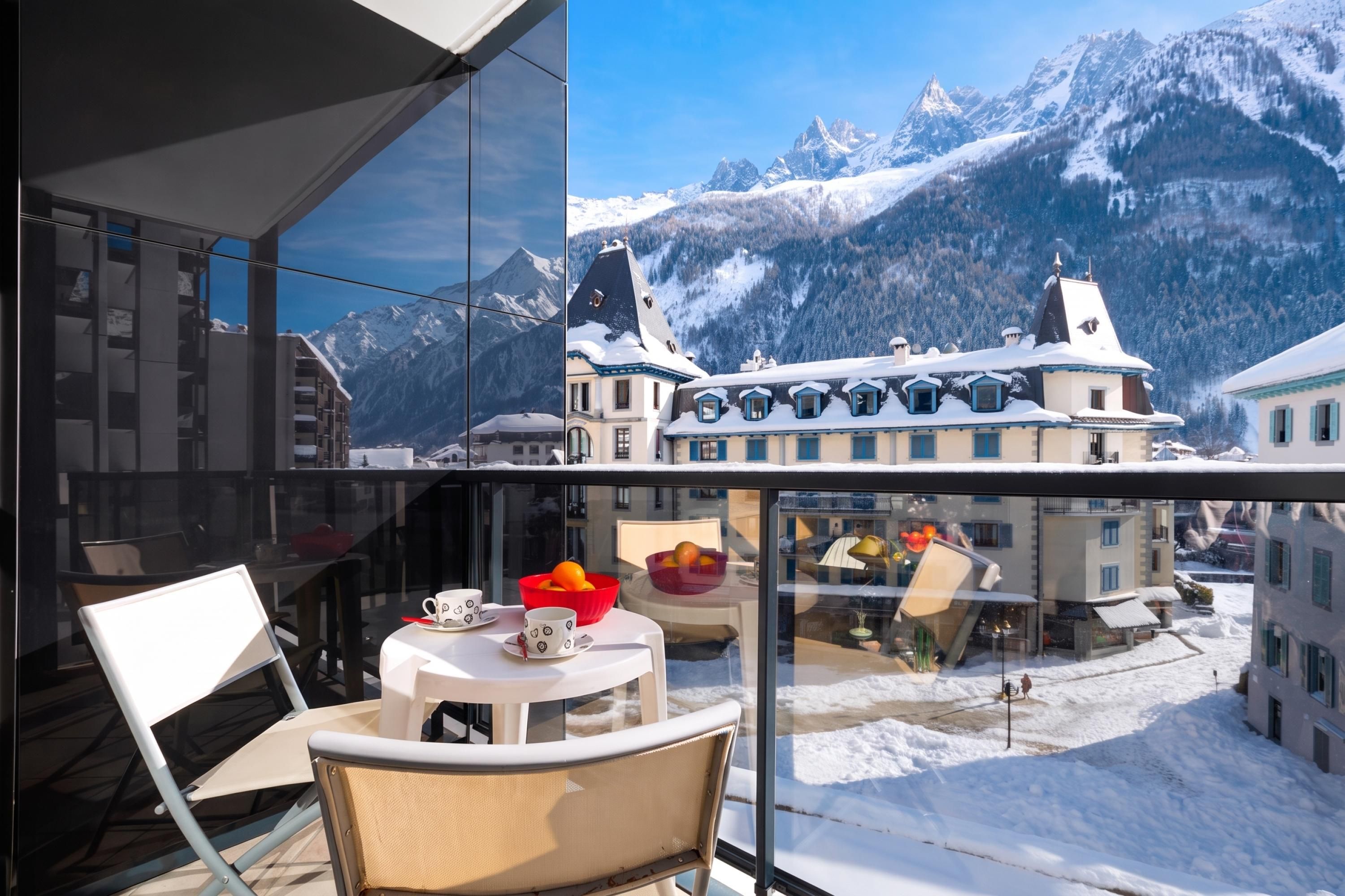 A balcony with a table, chairs, and a view of snow-covered mountains and buildings.