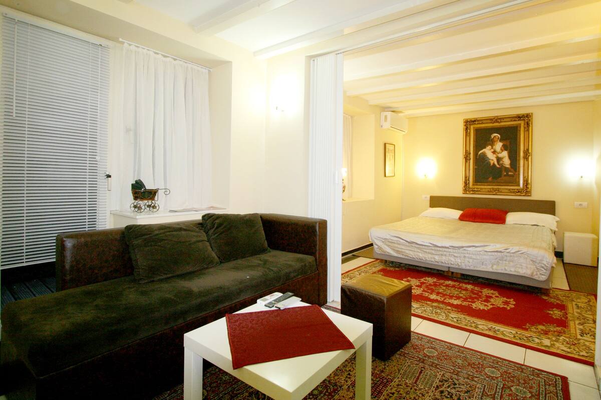 Ein Zimmer mit einer Couch, einem kleinen Tisch, einem Bett mit rotem Kopfkissen und einem Bild an der Wand.