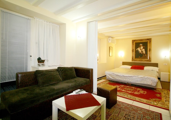 A room with a sofa, a small table, a bed with a red pillow, and a painting on the wall.
