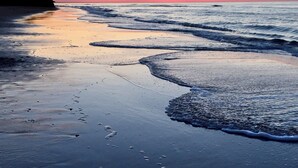 Une plage où les vagues viennent doucement lécher le rivage, un ciel dégagé et, au loin, la silhouette d'une falaise.