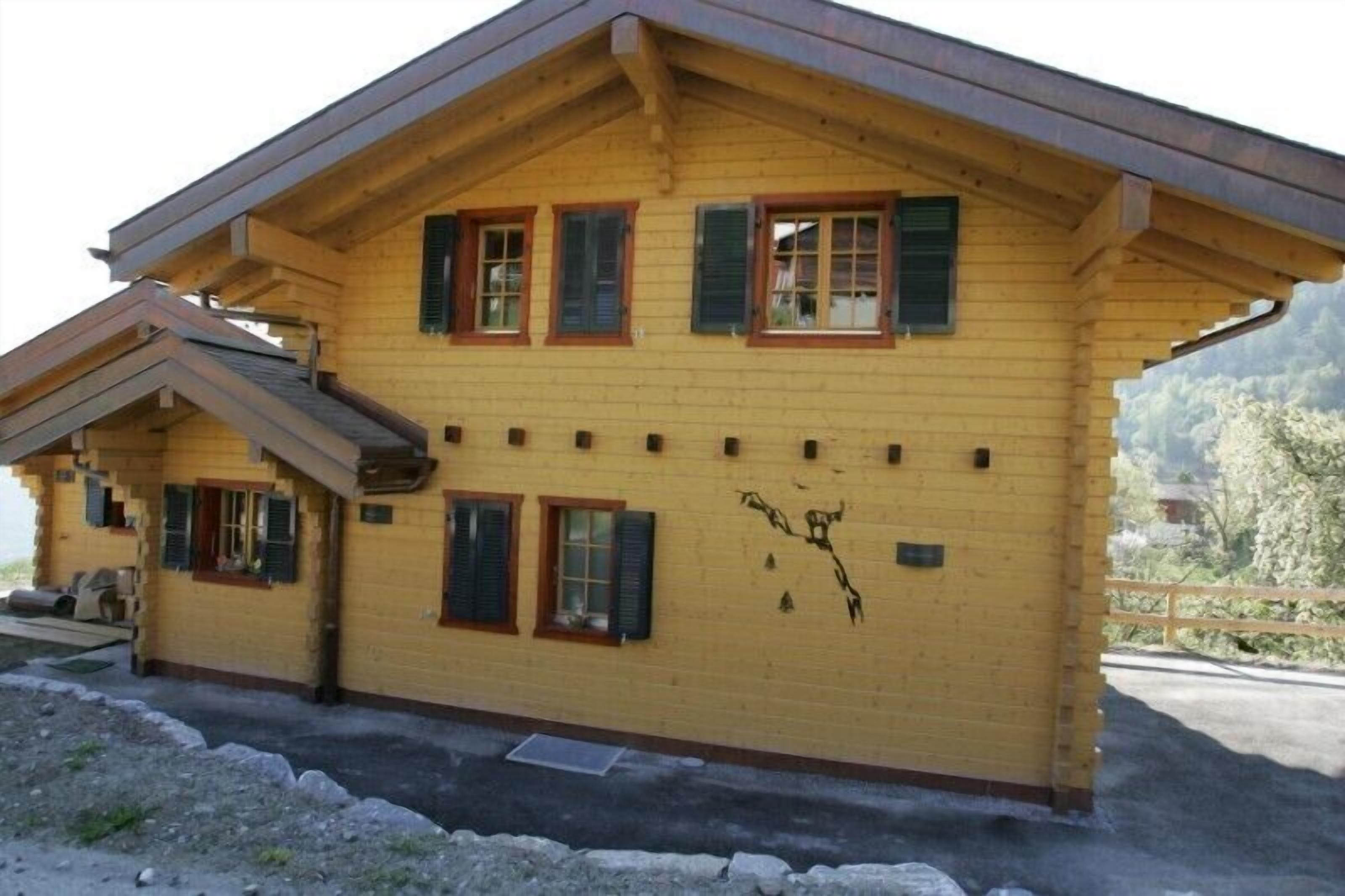 A two-story wooden house with a sloped roof, multiple windows with shutters, and a small balcony.