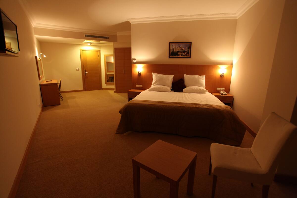 A hotel room with a large bed, bedside tables, a chair, a small table, and a painting on the wall.