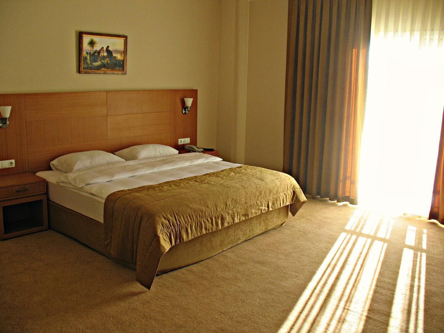 A hotel room with a large bed, wooden headboard, bedside tables, a painting on the wall, and a window with curtains.
